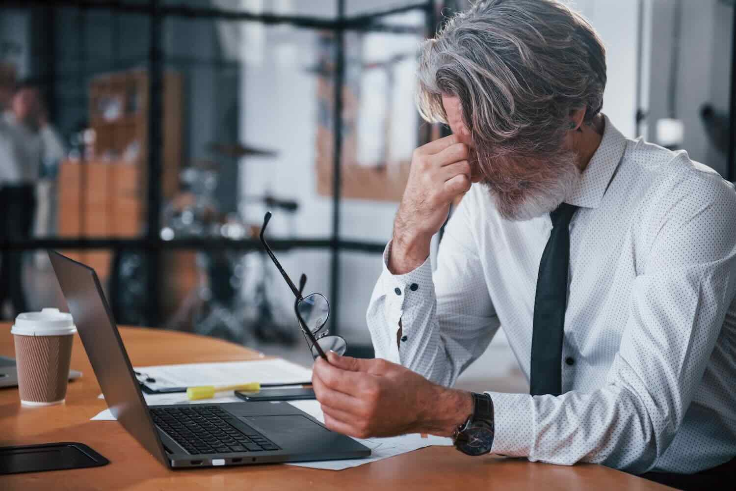 Tired mature businessman with grey hair and beard in formal clothes is in the office.