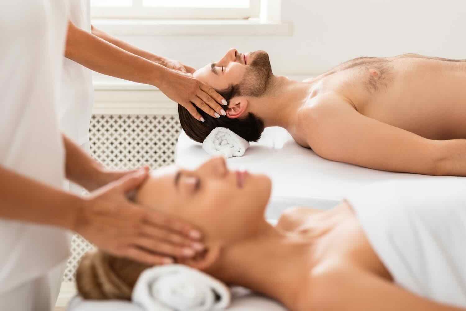 Relaxed Couple Receiving Head Massage Together Lying On Beds In Beauty Salon, With Eyes Closed, Focus On Husband. Beauty Treatment, Wellness And Relaxation. Date In Spa Concept